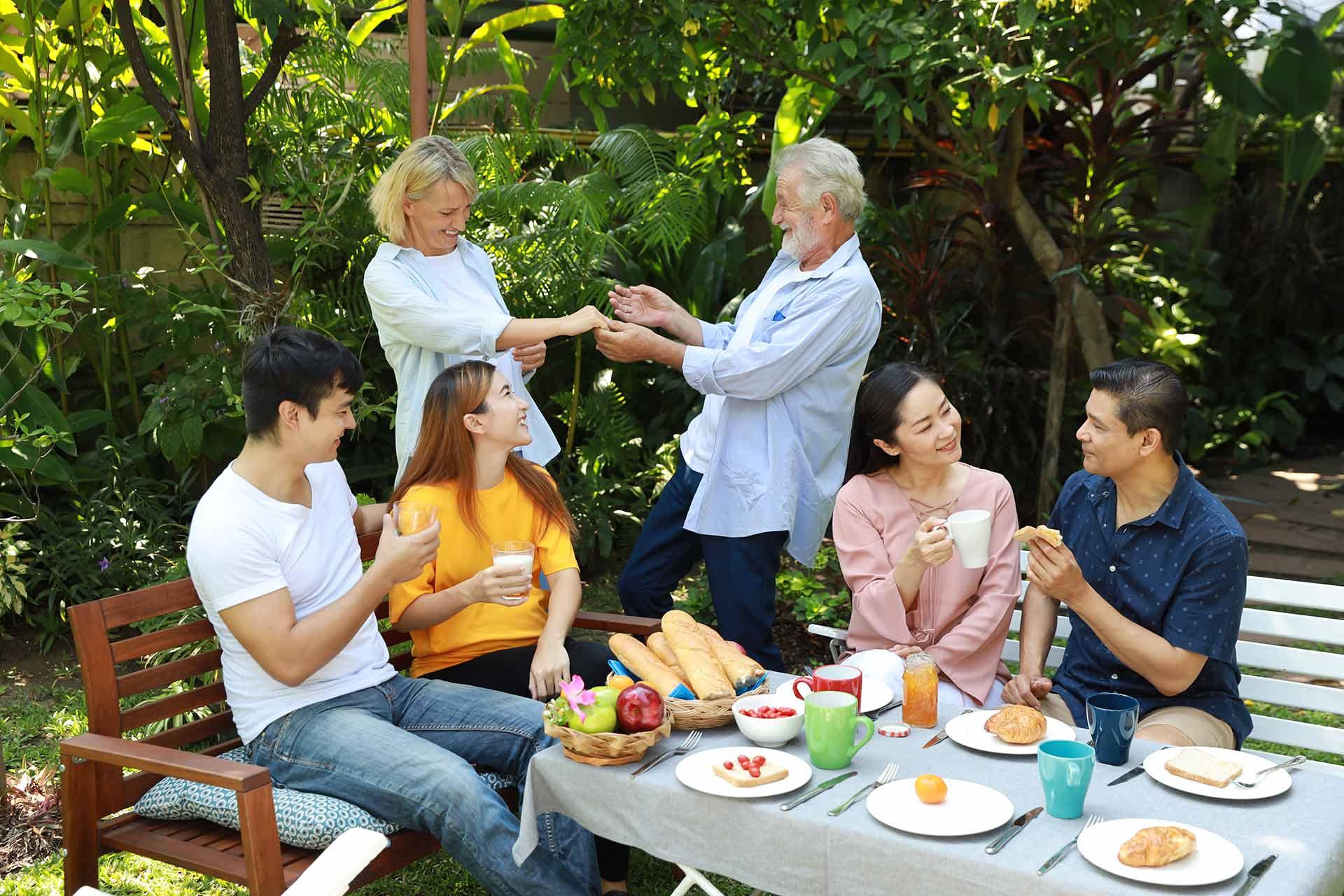 Happy Multiethnic Family Standing and Sitting at a Breakfast Table in Backyard Outdoor on Sunny Day With Smiling Face